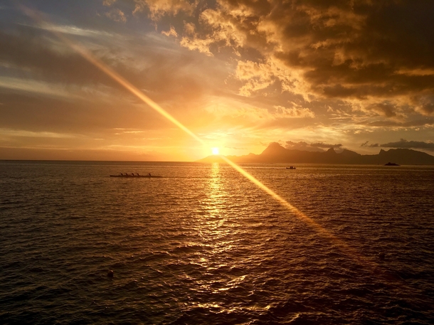 A sunset on Tahiti looking towards Moorea as a traditional Polynesian canoe rows by 