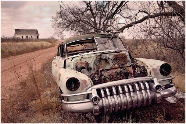 An abandoned car all torn up with bullet holes in the windshield and a boarded up old church in the background  by Evan Cohen